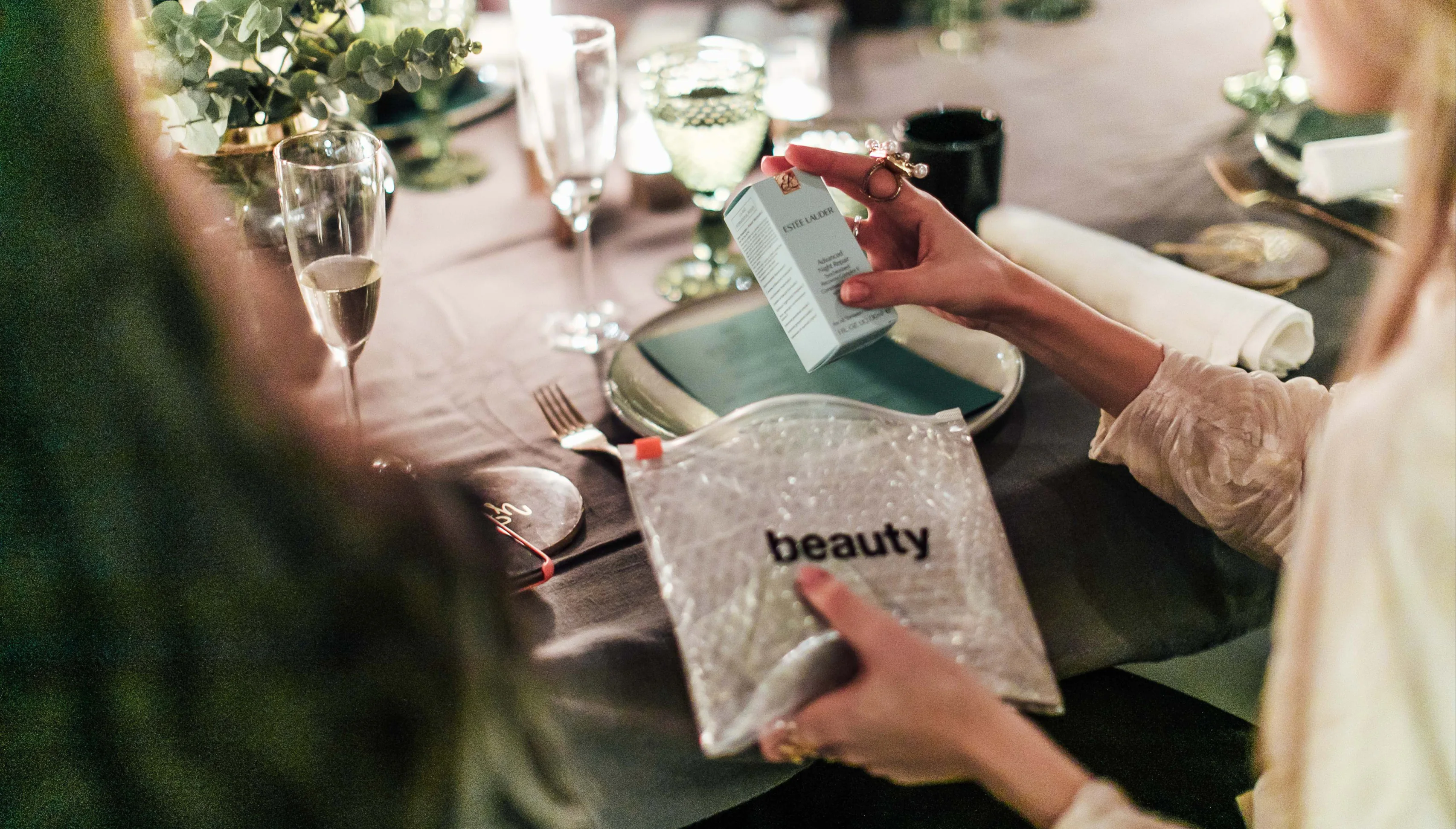 Hands holding a skincare box and a clear bag marked beauty at a dinner table